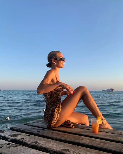 A woman in a leopard print dress sitting on a dock with a drink.