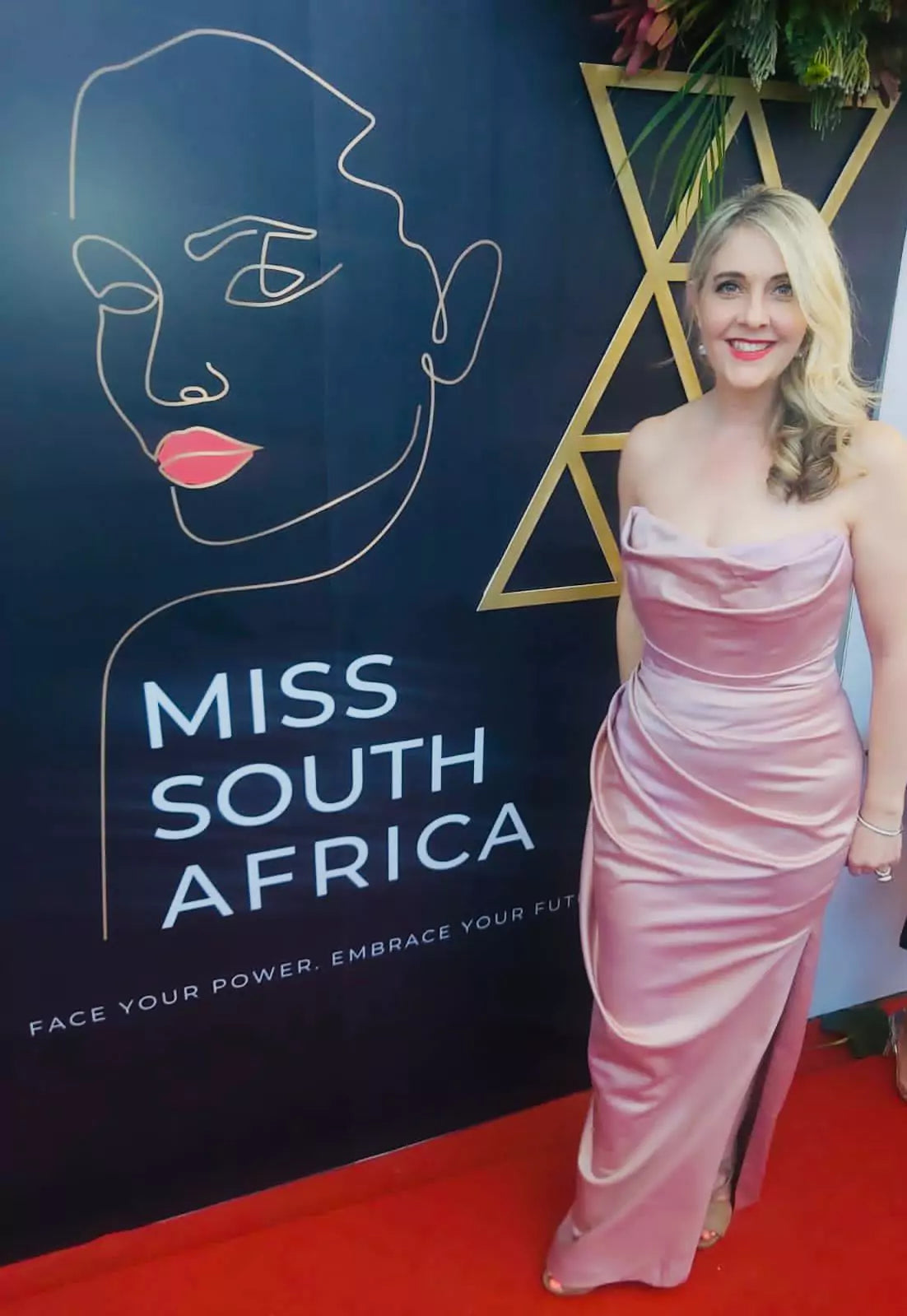 A woman in a pink dress standing in front of a miss south africa sign.
