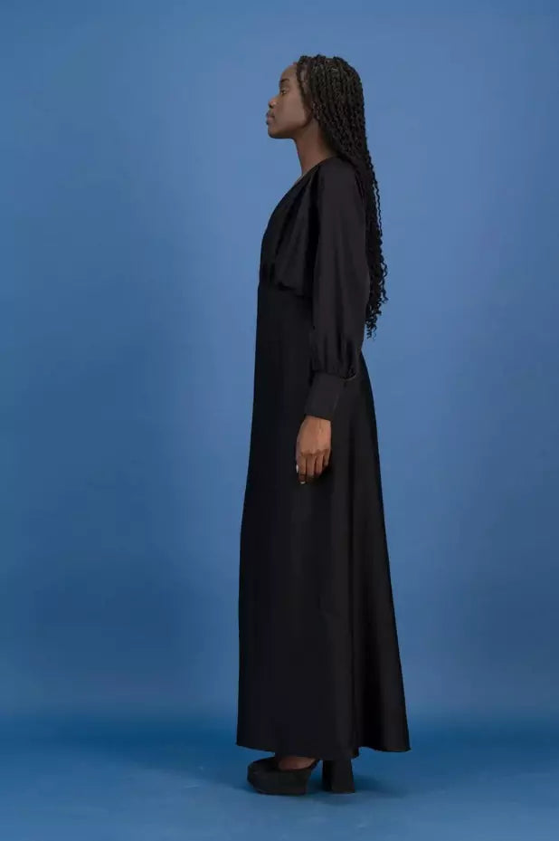 A woman in a black long dress standing in front of a blue background.