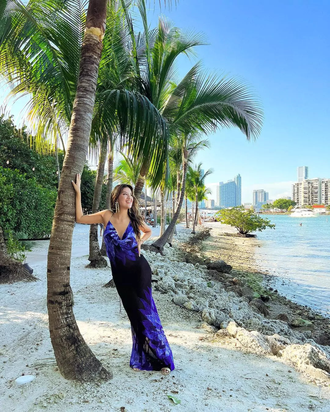 A woman in a purple dress leans on a palm tree
